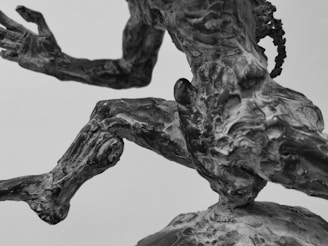 A close-up view of a metal sculpture depicting a human figure in motion. The figure's body appears muscular and detailed, with an expressive posture suggesting movement or action. The sculpture's texture is rough and metallic.
