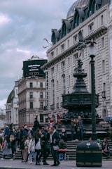 A bustling city street scene with historical architecture, featuring a crowd of people walking and gathering around a large ornate fountain. The buildings have classic European design with white stone facades and multiple stories. A digital billboard is visible in the background.