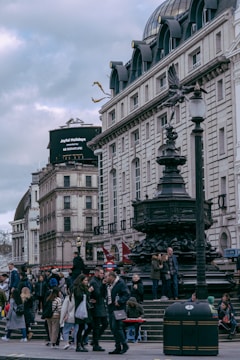 A bustling city street scene with historical architecture, featuring a crowd of people walking and gathering around a large ornate fountain. The buildings have classic European design with white stone facades and multiple stories. A digital billboard is visible in the background.