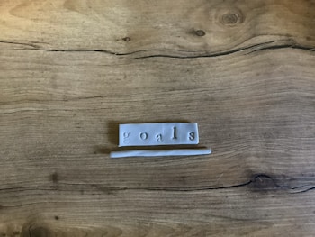 A piece of clay or similar material is shaped into a rectangular slab with the word 'goals' imprinted on it. The slab is placed on a wooden surface with natural grain patterns visible.