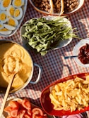 A table covered with a red and white checkered cloth is adorned with a variety of food. A dish contains sliced hard-boiled eggs, revealing their bright yellow yolks. Fresh green leafy vegetables are arranged on an oval platter. A round pan contains a creamy yellow dish, likely polenta or another grain-based meal. A red bowl is filled with crispy golden fries. Beside them are sliced fresh tomatoes. Nuts or dried fruits fill another sectioned dish. Background elements include a spoon and a small plate with a reddish condiment.