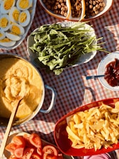 A table covered with a red and white checkered cloth is adorned with a variety of food. A dish contains sliced hard-boiled eggs, revealing their bright yellow yolks. Fresh green leafy vegetables are arranged on an oval platter. A round pan contains a creamy yellow dish, likely polenta or another grain-based meal. A red bowl is filled with crispy golden fries. Beside them are sliced fresh tomatoes. Nuts or dried fruits fill another sectioned dish. Background elements include a spoon and a small plate with a reddish condiment.