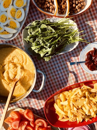 A table covered with a red and white checkered cloth is adorned with a variety of food. A dish contains sliced hard-boiled eggs, revealing their bright yellow yolks. Fresh green leafy vegetables are arranged on an oval platter. A round pan contains a creamy yellow dish, likely polenta or another grain-based meal. A red bowl is filled with crispy golden fries. Beside them are sliced fresh tomatoes. Nuts or dried fruits fill another sectioned dish. Background elements include a spoon and a small plate with a reddish condiment.