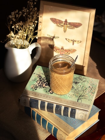 A moody still life of a jar of homemade kimchi resting on a worn wooden table surrounded by vintage books and dried flowers.