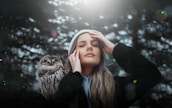 A person with closed eyes is gently touching their face, standing next to an owl in a serene forest setting. Soft sunlight filters through the trees, casting a peaceful glow.