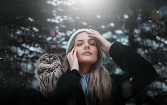A person with closed eyes is gently touching their face, standing next to an owl in a serene forest setting. Soft sunlight filters through the trees, casting a peaceful glow.