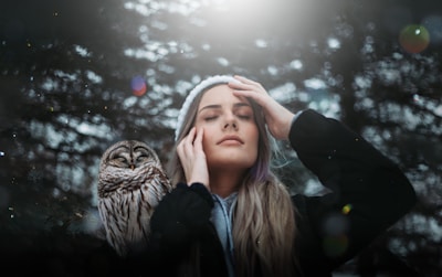 A person with closed eyes is gently touching their face, standing next to an owl in a serene forest setting. Soft sunlight filters through the trees, casting a peaceful glow.