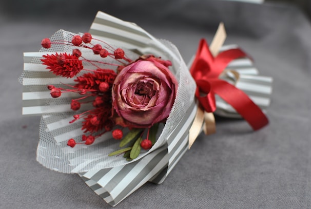 A bouquet consisting of a central pink rose surrounded by red dried flowers and green foliage. It is wrapped in striped paper with a decorative red ribbon tied around it, set on a soft gray background.