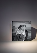 A framed black and white photograph features a couple dressed in wedding attire. The bride is holding a bouquet of flowers and looking content as the groom kisses her on the cheek. The image captures a romantic moment with a scenic beach backdrop visible in the background.
