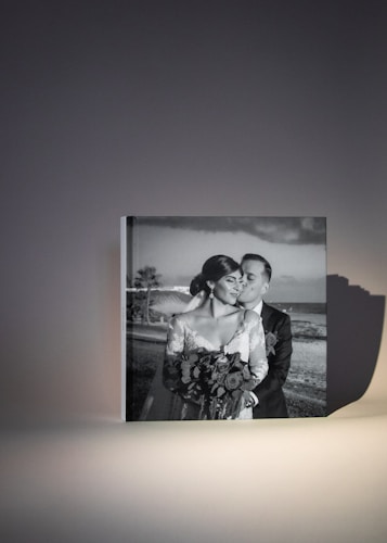 A framed black and white photograph features a couple dressed in wedding attire. The bride is holding a bouquet of flowers and looking content as the groom kisses her on the cheek. The image captures a romantic moment with a scenic beach backdrop visible in the background.