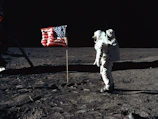 Silhouetted astronaut planting a flag on the lunar surface under a black sky.