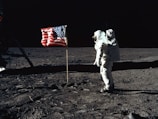 An astronaut stands on the surface of the moon near an American flag. The lunar landscape is barren and desolate, with a dark sky in the background. Footprints are visible on the dusty surface.