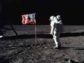 An astronaut stands on the surface of the moon near an American flag. The lunar landscape is barren and desolate, with a dark sky in the background. Footprints are visible on the dusty surface.