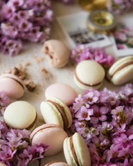 Macarons are arranged amidst a bed of vibrant pink flowers. The pastries vary in color, displaying soft pastels such as pink and cream with chocolate filling. In the background, blurred photographs and a decorative jar provide a vintage, artistic touch.