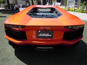 A bright orange Lamborghini with sleek lines and an aggressive rear design is parked on a grassy area. The car features a luxurious and aerodynamic structure, highlighting its sporty appearance.