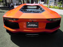 A bright orange Lamborghini with sleek lines and an aggressive rear design is parked on a grassy area. The car features a luxurious and aerodynamic structure, highlighting its sporty appearance.