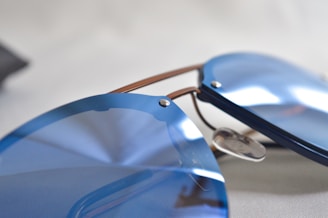 A close-up of sleek, modern sunglasses with reflective, blue-tinted lenses. The frames are minimalist, with subtle metallic accents, lying on a neutral background.