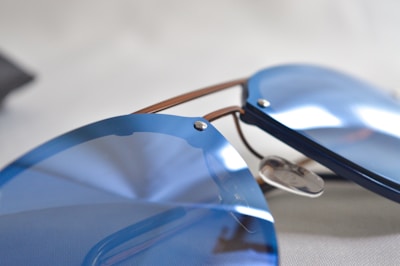 A close-up of sleek, modern sunglasses with reflective, blue-tinted lenses. The frames are minimalist, with subtle metallic accents, lying on a neutral background.
