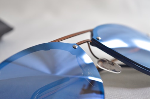 A close-up of sleek, modern sunglasses with reflective, blue-tinted lenses. The frames are minimalist, with subtle metallic accents, lying on a neutral background.