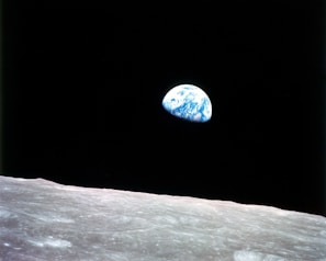 A view from the lunar surface shows Earth rising above the horizon, with a dark, expansive space in the background. The foreground is dominated by the barren, gray lunar landscape, while Earth appears as a distant, vibrant blue and white sphere.