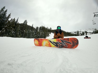 A person wearing winter gear sits on a snowy slope with a colorful snowboard featuring a fiery design. Coniferous trees line the side of the slope, and a partially cloudy sky looms overhead. A ski lift cable can be seen in the background.
