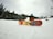 A person wearing winter gear sits on a snowy slope with a colorful snowboard featuring a fiery design. Coniferous trees line the side of the slope, and a partially cloudy sky looms overhead. A ski lift cable can be seen in the background.