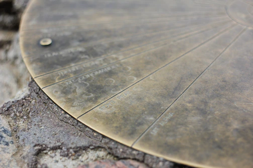 A close-up of a weathered metallic disk on a rocky surface, featuring engraved lines and text. The text includes names like 'BOYS PEAK' and 'MT. LIPIAH'. The disk appears to be part of a larger topographical or navigational instrument.