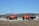 Two large, bright orange trucks with open frameworks on their beds are parked on a gravel area enclosed by low rope barriers. The trucks are designed for rugged terrain with large tires and elevated chassis. In the background, there are leafless trees and a small building with a pointed roof.