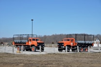 Two large, bright orange trucks with open frameworks on their beds are parked on a gravel area enclosed by low rope barriers. The trucks are designed for rugged terrain with large tires and elevated chassis. In the background, there are leafless trees and a small building with a pointed roof.