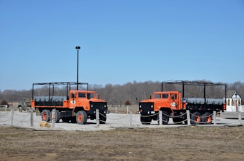 Two large, bright orange trucks with open frameworks on their beds are parked on a gravel area enclosed by low rope barriers. The trucks are designed for rugged terrain with large tires and elevated chassis. In the background, there are leafless trees and a small building with a pointed roof.