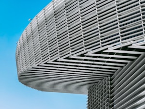 The image features the side view of a modern architectural structure with a series of parallel, horizontal slats or louvers. These create a dynamic pattern against the clear blue sky, giving a sense of movement and depth. The building appears to be made of a metallic material, reflecting light subtly.