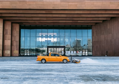 A yellow truck fitted with a snow plow is clearing snow in front of a large modern building with reflective windows. The facade of the building has a contemporary design with brown cladding and a series of vertical columns.