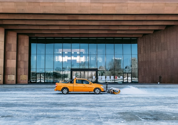 A yellow truck fitted with a snow plow is clearing snow in front of a large modern building with reflective windows. The facade of the building has a contemporary design with brown cladding and a series of vertical columns.