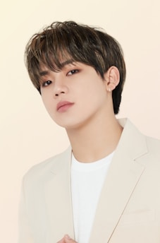 A person with short, dark hair is wearing a light-colored blazer over a white shirt. Their head is slightly tilted, and they have a thoughtful expression.