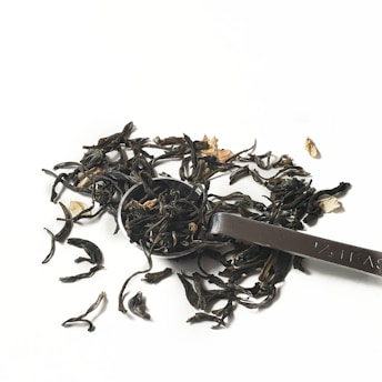 A metal teaspoon partially filled with loose-leaf tea is accompanied by scattered tea leaves on a white background. The tea appears dark with a few floral elements mixed in.