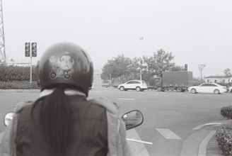 A black and white photograph capturing a person wearing a helmet and jacket, waiting at a traffic light. The view is from behind the person, looking toward a street intersection where several vehicles, including cars and a truck, are passing by. The environment features trees and street signs.