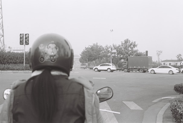 A black and white photograph capturing a person wearing a helmet and jacket, waiting at a traffic light. The view is from behind the person, looking toward a street intersection where several vehicles, including cars and a truck, are passing by. The environment features trees and street signs.