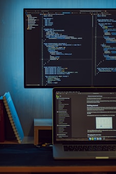 A monitor displaying lines of code in a text editor interface, placed above a laptop showing a text document. The setup is on a wooden desk with books and notebooks on the left side. The overall environment is minimalist and focused, suggesting a workspace for programming or writing.