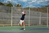 A person in athletic attire is playing tennis on an outdoor court. The player is preparing to hit a tennis ball with a racquet, mid-motion during a serve or a powerful shot. The court is surrounded by a wire fence, and the background shows a clear blue sky with scattered clouds.