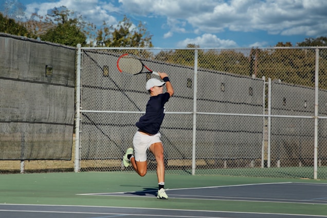 A person in athletic attire is playing tennis on an outdoor court. The player is preparing to hit a tennis ball with a racquet, mid-motion during a serve or a powerful shot. The court is surrounded by a wire fence, and the background shows a clear blue sky with scattered clouds.