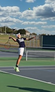 A tennis player is captured mid-air while serving at an outdoor tennis court. The player is wearing a navy blue shirt with 'Keller Tennis' printed on it, white shorts, and bright green sports shoes. A tennis racket is in the player's right hand, while the tennis ball is in the air above. The background includes a chain-link fence, a field, trees, and a partly cloudy sky.