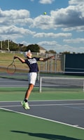 A tennis player is captured mid-air while serving at an outdoor tennis court. The player is wearing a navy blue shirt with 'Keller Tennis' printed on it, white shorts, and bright green sports shoes. A tennis racket is in the player's right hand, while the tennis ball is in the air above. The background includes a chain-link fence, a field, trees, and a partly cloudy sky.