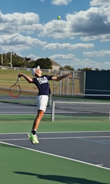 A tennis player is captured mid-air while serving at an outdoor tennis court. The player is wearing a navy blue shirt with 'Keller Tennis' printed on it, white shorts, and bright green sports shoes. A tennis racket is in the player's right hand, while the tennis ball is in the air above. The background includes a chain-link fence, a field, trees, and a partly cloudy sky.