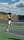 A tennis player is captured mid-air while serving at an outdoor tennis court. The player is wearing a navy blue shirt with 'Keller Tennis' printed on it, white shorts, and bright green sports shoes. A tennis racket is in the player's right hand, while the tennis ball is in the air above. The background includes a chain-link fence, a field, trees, and a partly cloudy sky.