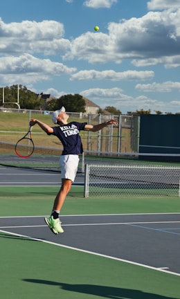 A tennis player is captured mid-air while serving at an outdoor tennis court. The player is wearing a navy blue shirt with 'Keller Tennis' printed on it, white shorts, and bright green sports shoes. A tennis racket is in the player's right hand, while the tennis ball is in the air above. The background includes a chain-link fence, a field, trees, and a partly cloudy sky.