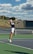 A tennis player is captured mid-air while serving at an outdoor tennis court. The player is wearing a navy blue shirt with 'Keller Tennis' printed on it, white shorts, and bright green sports shoes. A tennis racket is in the player's right hand, while the tennis ball is in the air above. The background includes a chain-link fence, a field, trees, and a partly cloudy sky.