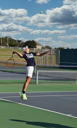 A tennis player is captured mid-air while serving at an outdoor tennis court. The player is wearing a navy blue shirt with 'Keller Tennis' printed on it, white shorts, and bright green sports shoes. A tennis racket is in the player's right hand, while the tennis ball is in the air above. The background includes a chain-link fence, a field, trees, and a partly cloudy sky.