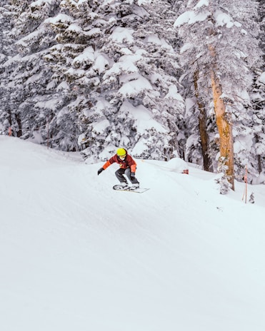 A snowboarder is mid-air, performing a jump on a snowy slope surrounded by snow-covered pine trees. The snowboarder is wearing a helmet and winter sports gear, including a bright yellow helmet that stands out against the white snow.