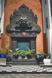 An intricately carved black stone facade featuring a prominent central figure above a doorway, set against a red brick background. Potted plants line the entrance, and stone elephant statues flank the steps leading up to the checkered tile floor.