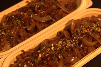 Two rectangular containers are filled with a savory dish consisting of caramelized onions and what appears to be slices of beef or a similar protein, garnished with herbs and small bits of seasoning. The food has a glossy appearance due to the sauce, and the containers are placed side by side.